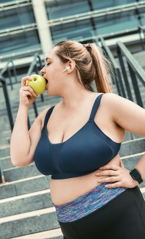 Healthy food. Plus size woman in sport clothing eating green apple while standing on stairs outside